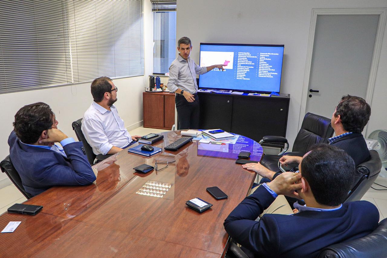 Foto da reunião com o presidente Gilson Santos em pé apontando para uma televisão com um mapa